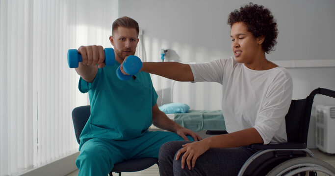 Doctor helping patient in wheelchair with dumbbells doing rehabilitation exercise in medical center
