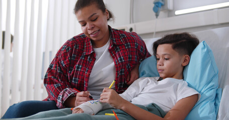 Black mother visiting son lying with broken arm in bed in hospital ward