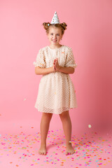 Happy little girl in a birthday cap blows off her palms multicolored confetti on a pink background in the studio