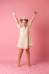 Happy little girl in a birthday cap blows off her palms multicolored confetti on a pink background in the studio