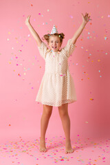 Happy little girl in a birthday cap blows off her palms multicolored confetti on a pink background in the studio