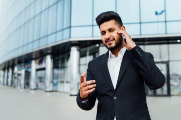 Portrait of a smiling serious Indian man talking on wireless headphones and gesturing to the camera, place for text