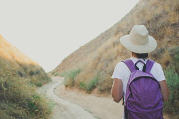 Fototapeta na wymiar Back view of woman hiker traveler walking on path in canyon gorge of national landscape park,wearing purple backpack, summer straw hat and white t-shirt.Active travel and adventure concept.Copy space