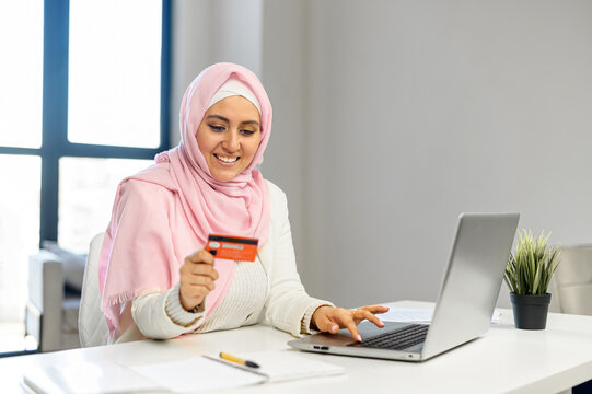 Young Muslim Arab mixed-race businesswoman in hijab sitting at the desk and entering typing credit card number on laptop, feeling excited to purchase items from an online store, order food, pay bills