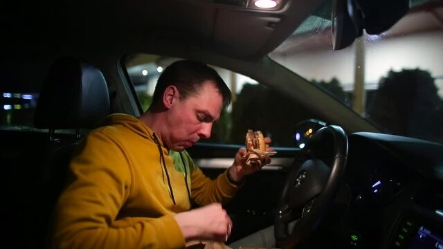 Hungry Man Eating Snacks And Burger In The Car. Satisfy Your Hunger On The Road. Dine In The Evening Car. Dark Background.