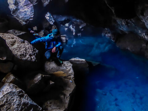 A young man wearing blue jacket squats at the rock in  Grjótagjá Cave, looking down at the crystal blue water. Famous Game of thrones location. The water in this cave is full of minerals, it's hot pot