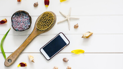 Smartphone mockup on white wooden background with leaves, flower petals, cosmetic bottles, shells, oils, salt, massage brush, copy space, flat lay, top view