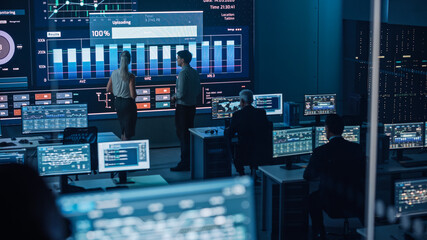 Two Software Engineers Talking Next to a Big Screen in a Modern Monitoring Office with Live Analysis Feed with Charts. Monitoring Room Big Data Scientists and Managers Sit in Front of Computers.