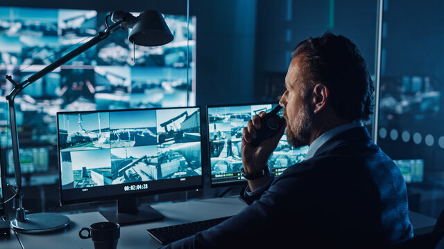 Male Officer Works On A Computer With Surveillance CCTV Video In A Harbour Monitoring Center With Multiple Cameras On A Big Digital Screen. Employee Uses Radio To Give An Order Or Report.