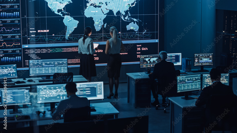 Poster Two Software Engineers Talking Next to a Big Screen in a Modern Monitoring Office with Live Analysis Feed with Charts. Monitoring Room Big Data Scientists and Managers Sit in Front of Computers.