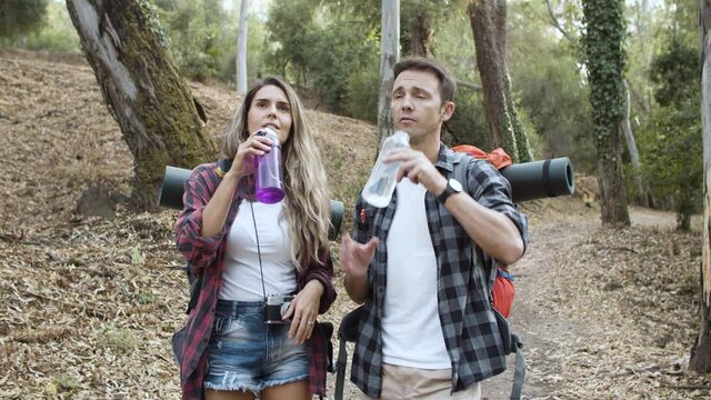 Couple of backpackers drinking fresh water on their way, walking on forest path for camping, enjoying outdoor leisure and healthy recreation. Adventure travel and active lifestyle concept