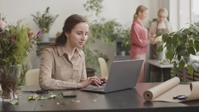 Medium close-up of young Caucasian woman sitting by desk in flower shop, using laptop computer, smiling, then calling for female colleagues working on background
