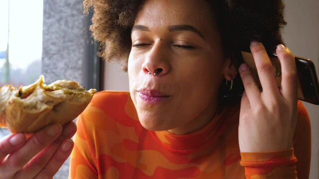 Young beautiful multiethnic woman indoors restaurant biting hamburger using smartphone - hunger, delicious, eating concept