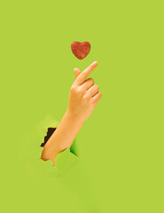 K pop concept. A girl hand showing fingers heart gesture. Red glittery heart above. Bright green in background.