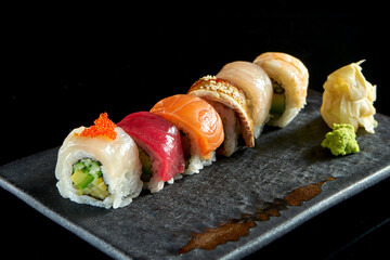 Rainbow sushi roll with different types of seafood: salmon, eel, shrimp, scallop, tuna and avocado, served on a ceramic plate with ginger and wasabi. Isolated on a black background. Japanese food