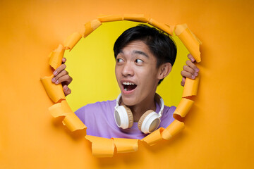 Young Asian teenager man pointing a copy space, poses through torn yellow paper hole, wearing purple t-shirt with headphone