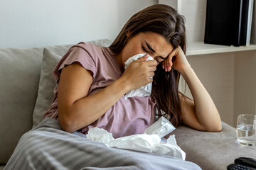 Shot of a frustrated woman using a tissue to sneeze in while being seated at home. Photo of sneezing woman in paper tissue. Attractive young girl feeling ill and blowing her nose with a tissue at home