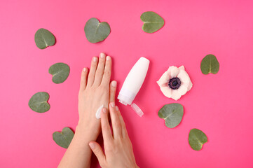 Woman applyiing cosmetic cream on her hands on pink background with flower and leaves. Home spa, beauty and treatment concept. Flat lay, top view