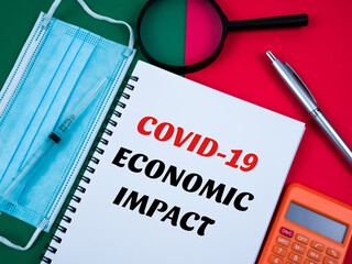 Top view of facemask, pen, calculator, syringe, magnifying glass, and notebook written a "COVID-19 ECONOMIC IMPACT" isolated on a green and red background. Business concept.