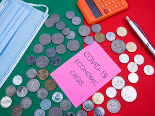 Top view of calculator, pen, facemask, coins and pink paper written a "COVID-19 ECONOMIC CRISIS" isolated on a green and red background. Business concept.