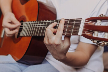 Fototapeta na wymiar Close up of hand of young woman in the white t-shirt and blue jeans playing acoustic guitar. Girl picks a barre chord clamping frets on the fretboard