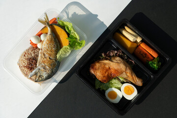 Top view of deep fried mackerel fish and rice with roasted grill chicken and egg in side the contain box over black and white pattern background.