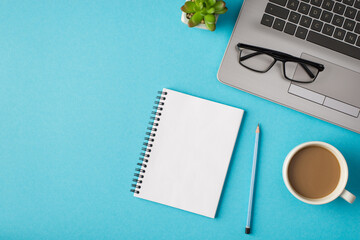 Top view photo of workplace with glasses on laptop pencil notebook plant and cup of coffee on isolated blue background with copyspace