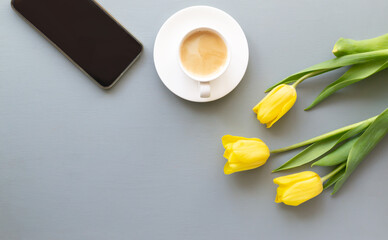Three yellow tulips, a cup of coffee, a smartphone lies on a gray background. Spring holidays concept. Top view. Copy space.