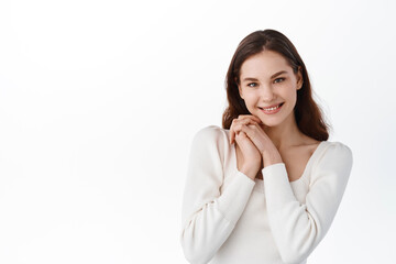 Fototapeta premium Cute young female model look heartfelt and pleased, holding hands near cheeks and smiling at camera, being praised, staring thankful and delighted, standing against white background