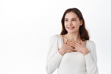 Pleased smiling woman keeps hands together on chest, expresses gratitude, looks aside, touched with...