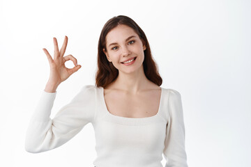Obraz premium Attractive woman in blouse showing OK sign and smiling satisfied, give positive reply, show alright agree sign, making okay and nod in approval, standing against white background. Excellent job