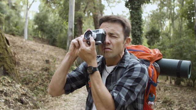 Backpacker guy with photo camera taking pictures of forest landscape, while walking on path, enjoying outdoor leisure time and healthy recreation. Adventure travel concept