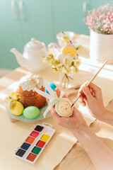 the hands of an unknown woman draw a bunny face on the egg. preparation for the holiday in the kitchen. on the table there are Easter attributes - Easter cake and colorful eggs.