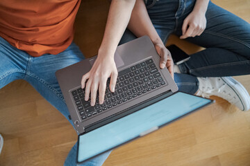 Young interracial couple sitting on the floor while using the laptop and consulting the mobile