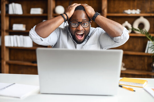 Shocked man is staring on the laptop screen, holding head and yelling happily, surprised African-American businessman received good news, won in lottery, male jobseeker get an unbelievable opportunity
