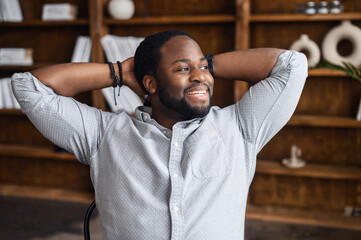 Carefree and successful African-American businessman taking a break on the workplace, a black guy in smart casual shirt leaned on the chair, put his hands behind his head, smiles