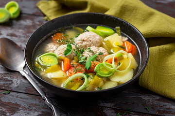 Rustic soup with vegetables and meatballs in a bowl on a dark wooden table. Soup with meatballs, leeks, potatoes, carrots and herbs. Tasty lunch or dinner rustic style.
