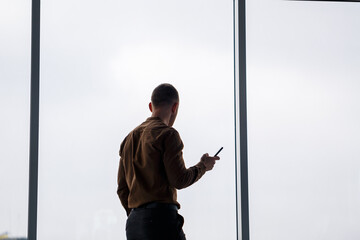 A young successful male businessman stands loosely near a large window and looks out over the city.