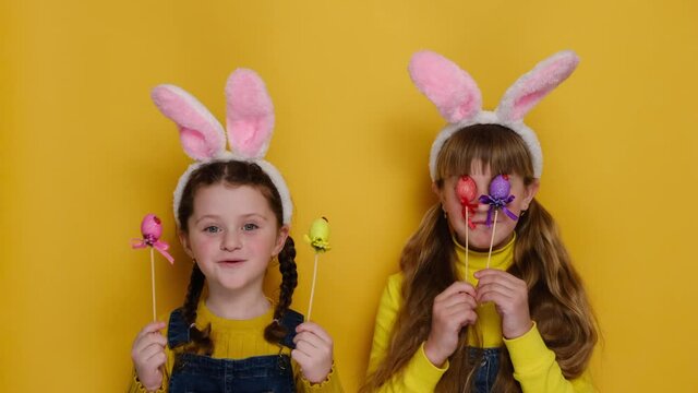 Funny cute child sisters in pink bunny ears studio isolated on yellow background looks positively at camera, holds and covers eyes with colored eggs. Childhood lifestyle and Happy Easter concept