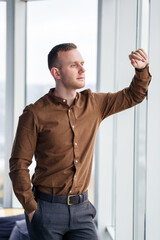 Fototapeta premium A young successful male businessman stands loosely near a large window and looks out over the city.