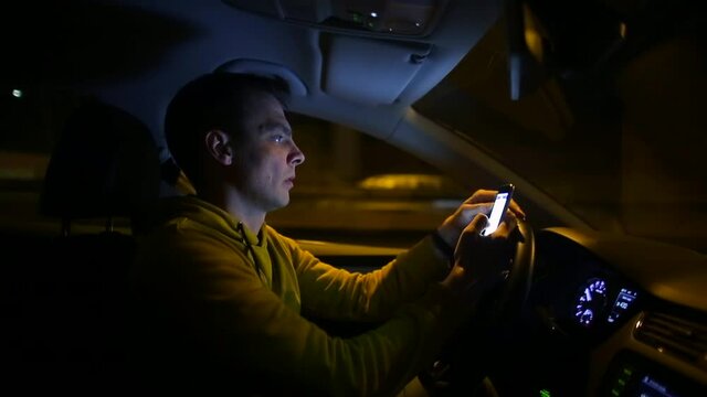 The male driver uses the phone while driving at night. He looks at the message along the way. Communication on the way. A road in bokeh is moving in the background.