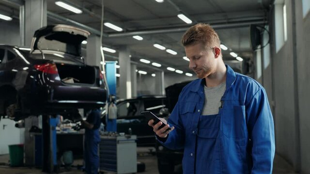 Handsome young man talking on the mobile phone and smiling while standing in workshop with car in the background mechanic