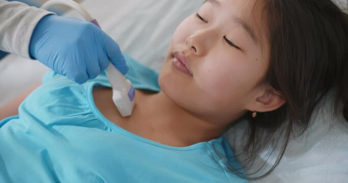 Preteen asian girl lying on bed while getting ultrasound of thyroid from doctor at hospital