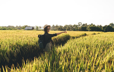 Naklejka na ściany i meble Portrait of cheerful female tourist with travel backpack pointing far away to rural landscape and smiling at camera, happy hipster girl in sunglasses enjoying solo travelling for visiting rice fields