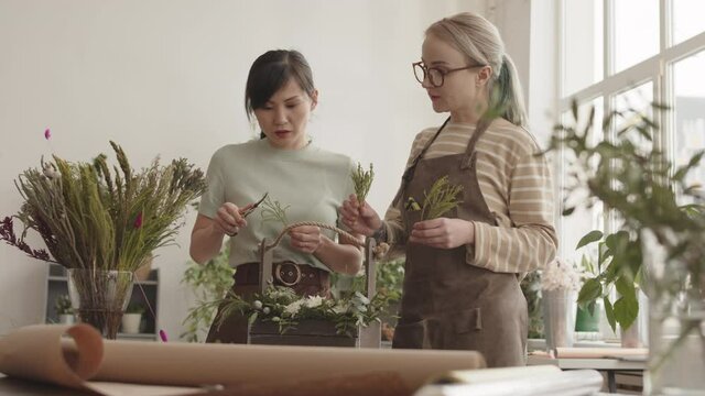 Medium long of blonde Caucasian and young Asian women standing in plant shop, talking, decorating wooden basket with flowers and greens