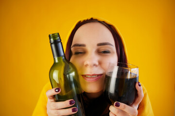 Relaxed young woman poses with bottle and glass of red wine on yellow background. Adult happy brunette presses alcohol to face.