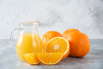 A glass pitcher of juice with fresh orange fruits on stone background