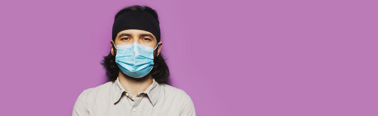 Studio portrait of young brunette guy, with long hair and protective medical face mask, on the background of purple wall.