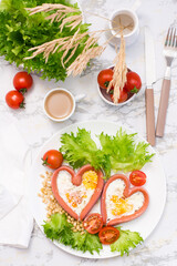 Fototapeta na wymiar Romantic breakfast. Fried eggs in heart shaped sausages, lettuce and cherry tomatoes on a plate on the table. Top and vertical view