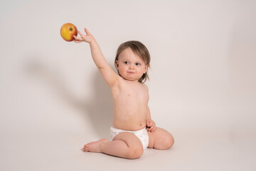 cute baby boy (one year old) sits in a diaper and throws a red fresh apple with his right hand, light background